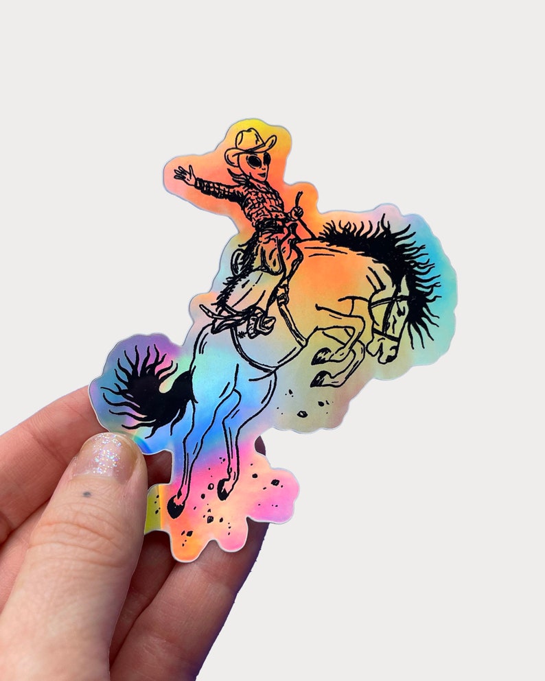 Peut inclure: Un autocollant holographique repr&eacute;sentant une illustration en noir et blanc d'un extraterrestre portant un chapeau de cowboy chevauchant un cheval cabr&eacute;.