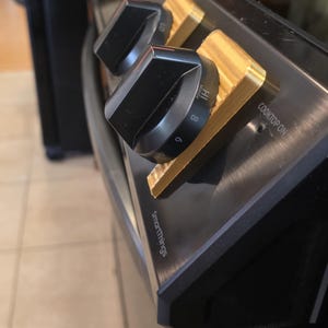 May include: A close-up of a black and gold stove top featuring four control knobs. The black knobs have white markings and are set on gold bases. The stove top displays the "SmartThings" logo and "Cooktop On" text.