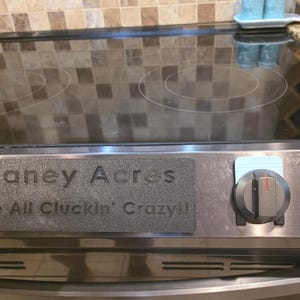 Puede incluir: Una cubierta rectangular negra texturizada para estufa con el texto en relieve Chaney Acres y We're All Cluckin' Crazy!! colocada en el panel de control de acero inoxidable de una estufa eléctrica con superficie de vidrio negro.