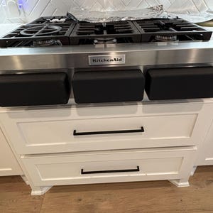 Puede incluir: Cocina de gas KitchenAid de acero inoxidable con rejillas y quemadores negros. La cocina tiene paneles de control negros y está instalada sobre gabinetes blancos con manijas negras. El salpicadero es blanco.