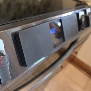 May include: A stainless steel oven control panel featuring a black rectangular display screen showing blue digital numbers. The panel includes a metallic control knob with a red indicator line on the left side and a smooth, dark finish.
