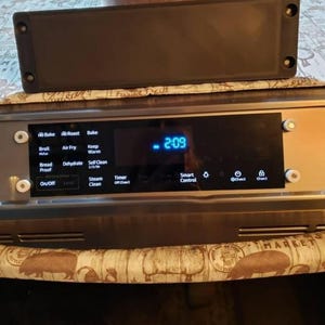 May include: A stainless steel oven control panel with a digital clock displaying 2:09. The panel features various function buttons like Bake, Broil, and Air Fry. A black rectangular cover sits above the panel.