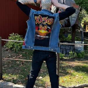 May include: A person wearing a blue denim vest with a Dio "The Last in Line" patch on the back. The person is wearing black jeans and white sneakers.