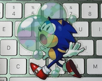 May include: A sticker of Sonic the Hedgehog, a blue anthropomorphic hedgehog, running across a computer keyboard. The sticker has a transparent bubble effect overlaying the keyboard keys. The keyboard is silver with white keys.
