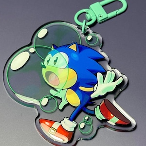 May include: A blue and yellow cartoon character, Sonic the Hedgehog, is running inside a clear acrylic keychain with a green metal clasp. The keychain is shaped like a bubble and has a green outline.