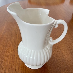 May include: A white ceramic jug with a fluted design and a handle. The jug is on a wooden table.