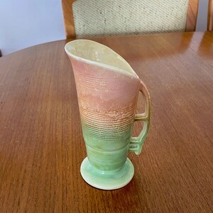 May include: A vintage art deco jug with a handle and a wide, curved spout. The jug features a colour gradient, transitioning from pink to orange, then to green. It sits on a small, round base and has a matching green handle.