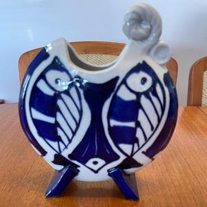 May include: A ceramic vase featuring a blue and white fish design. The vase has a rounded form, a handle, and is supported by three small feet. The fish design is symmetrical and stylized.