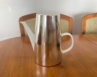 Op de afbeelding: Een zilveren theepot met een witte handgreep en tuit. De theepot staat op een houten tafel.