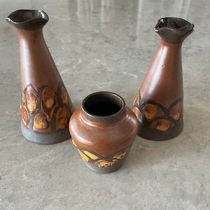 May include: Three ceramic vases in shades of brown and orange. Two tall, conical vases and one shorter, round vase. The vases have a textured design with a dark brown rim and base. The vases are likely for home decor.