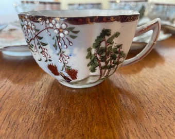 May include: A white porcelain teacup with a gold rim and floral and tree designs. The cup has a handle and is decorated with a delicate floral pattern and a scene of trees.