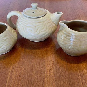 May include: A light beige ceramic tea set comprising a teapot with lid, a creamer jug, and a sugar bowl. Each piece features a textured, ridged design. The set is designed for serving tea.