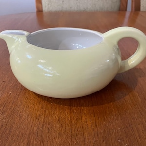 May include: A pale yellow ceramic creamer with a rounded body, a spout, and a loop handle. The interior is white. The creamer is on a wooden surface. Suitable for serving cream or milk.