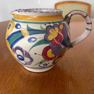 May include: A white ceramic jug with a floral design in shades of yellow, red, blue, and green. The jug has a pink handle with a silver trim.