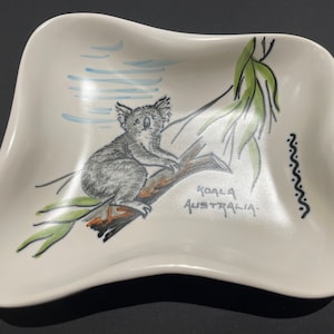 May include: A white ceramic dish with a whimsical hand-painted design of a koala bear sitting on a branch. The dish has a wavy edge and features the words "Koala Australia" in black script.
