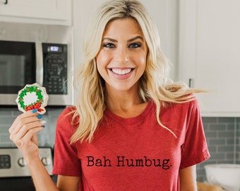 Könnte beinhalten: Eine Person trägt ein rotes T-Shirt mit dem Text "Bah Humbug" und hält einen Weihnachtskranz-Keks. Ein Teller mit dekorierten Weihnachtsplätzchen steht auf einer weißen Arbeitsplatte. Die Küche hat weiße Schränke und Edelstahlgeräte.