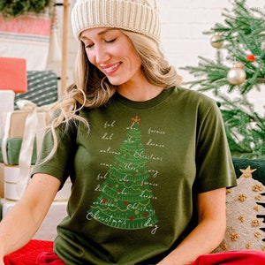 May include: An olive green t-shirt with a Christmas tree design and the words "ful, lal, welcome, Christmas day" in white script. The person is wearing a cream-colored knit hat and red sweatpants. A Christmas tree and wrapped gifts are in the background.