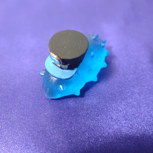 May include: A small, decorative bottle stopper featuring a blue, spiky base with a dark gray top and a light blue accent. The stopper has a small gold-colored detail and is set against a purple background.
