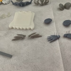May include: Assortment of grey plastic dollhouse furniture and accessories, including a small picture frame, a pair of wings, a pair of hands, and a pair of round decorations.