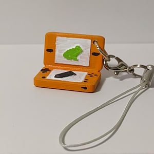 May include: Orange and white Nintendo DS keychain with a green frog on the screen and a black stylus. The keychain has a white string.