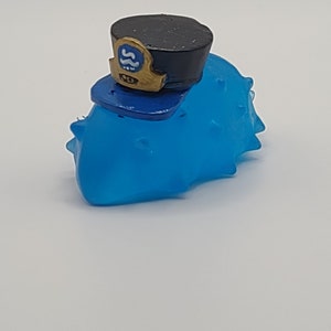 May include: A blue and black toy with a spiky, textured surface. The toy is wearing a black and gold hat with a blue and white emblem that says "S.S.D."