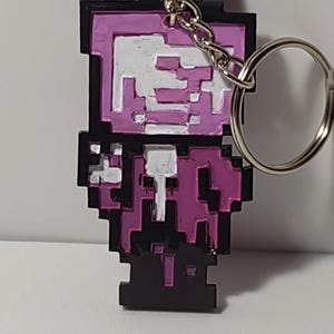 Puede incluir: Llavero pixelado con un diseño de robot morado y blanco, una cadena plateada y un anillo metálico. El robot tiene un contorno negro y una estética de videojuego retro. Un accesorio divertido.