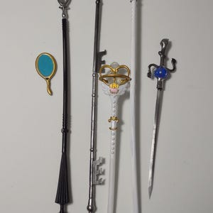 Puede incluir: Colección de accesorios de fantasía, que incluyen un bastón negro con un extremo en forma de abanico, un bastón plateado en forma de llave con un corazón rosa y un bastón blanco con una corona. También incluye un pequeño espejo y una espada.