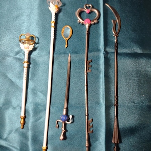 Puede incluir: Una colección de cinco objetos de fantasía en miniatura que incluye dos varitas blancas con detalles dorados, un pequeño espejo de mano ovalado con marco dorado, un bastón plateado en forma de llave con gemas rosas y una lanza metálica oscura.