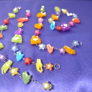 May include: A collection of colorful beaded jewelry, including necklaces and a bracelet, featuring pixelated star and frog shapes. The beads are in shades of yellow, orange, green, blue, purple, and red, strung on silver chains, set against a purple background.