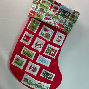 Op de afbeelding: Rode kerstsok met een Grinch-thema stofmanchet. De sok heeft 12 genummerde vakjes, elk met een ander Grinch-thema afbeelding. De manchet heeft de tekst "12 Days of Grinchmas" en verschillende Grinch-thema illustraties.
