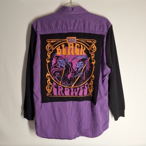May include: A long-sleeved shirt with a striking design. The back features a large black patch with the words "THE BLACK CROWES" in orange, along with an illustration of two grim reapers. The shirt combines purple pinstripes and black sleeves.