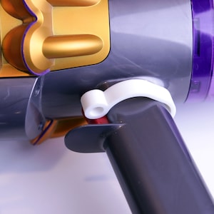 May include: A close-up of a gray and purple vacuum cleaner with a white plastic release latch. The vacuum cleaner has a gold-colored dustbin.