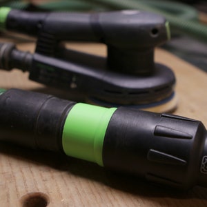 May include: A close-up of a black and green power sander with a green dust collection hose. The sander has a black handle and a green trigger. The hose is connected to the sander with a black and green connector.