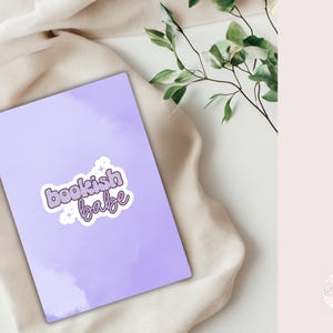 Puede incluir: Una tarjeta rectangular de color lavanda que presenta una pegatina blanca con el texto bookish babe en letras de burbuja moradas, acentuado por pequeños iconos de estrellas blancas. La tarjeta tiene una textura de acuarela suave.