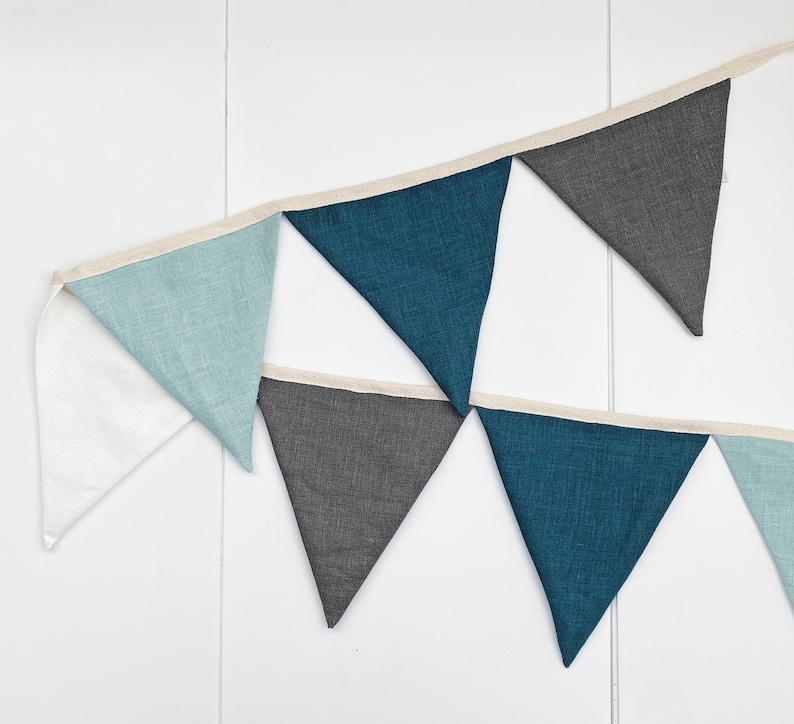 May include: A bunting banner with alternating blue, gray, and white triangular flags. The flags are made of a textured fabric and are connected by a white ribbon.