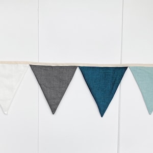 May include: A bunting banner with five triangular flags in white, gray, teal, and light blue colors. The flags are sewn together with a white string.