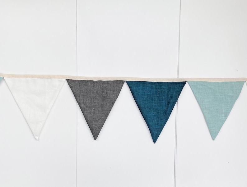May include: A bunting banner with five triangular flags in white, gray, teal, and light blue colors. The flags are sewn together with a white string.