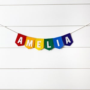 May include: A colorful felt banner spelling "AMELIA" in white letters. Each letter is on a different colored pennant: red, yellow, green, and blue. The banner is strung on a thin white string against a white background.