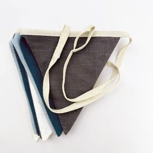 May include: A set of five fabric pennant flags in shades of blue, gray, and white. The flags are sewn together with a white cotton ribbon.