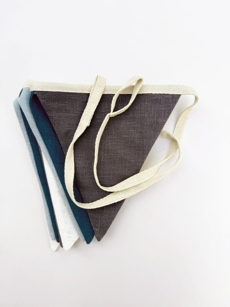 May include: A set of five fabric pennant flags in shades of blue, gray, and white. The flags are sewn together with a white cotton ribbon.