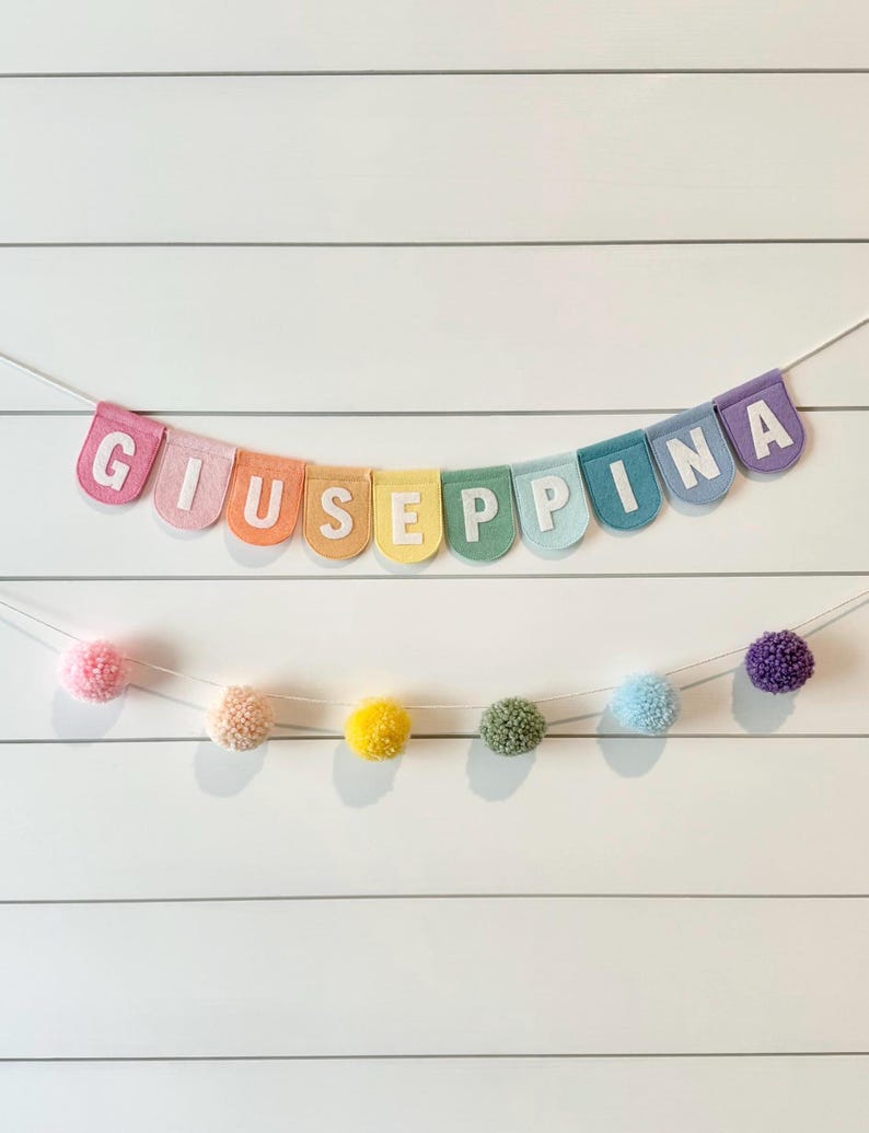 May include: A colorful banner spelling "GIUSEPPINA" in white letters, strung across a white background. The banner is composed of pastel-colored pennants. Below, a garland of pom-poms in various colors hangs, adding a playful touch.
