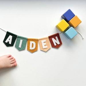 May include: A colorful felt banner spelling "AIDEN" with white letters on various colored pennants. The banner is strung on a white cord, and a cluster of wooden blocks in blue, yellow, and orange are nearby. A child's foot is visible in the corner.