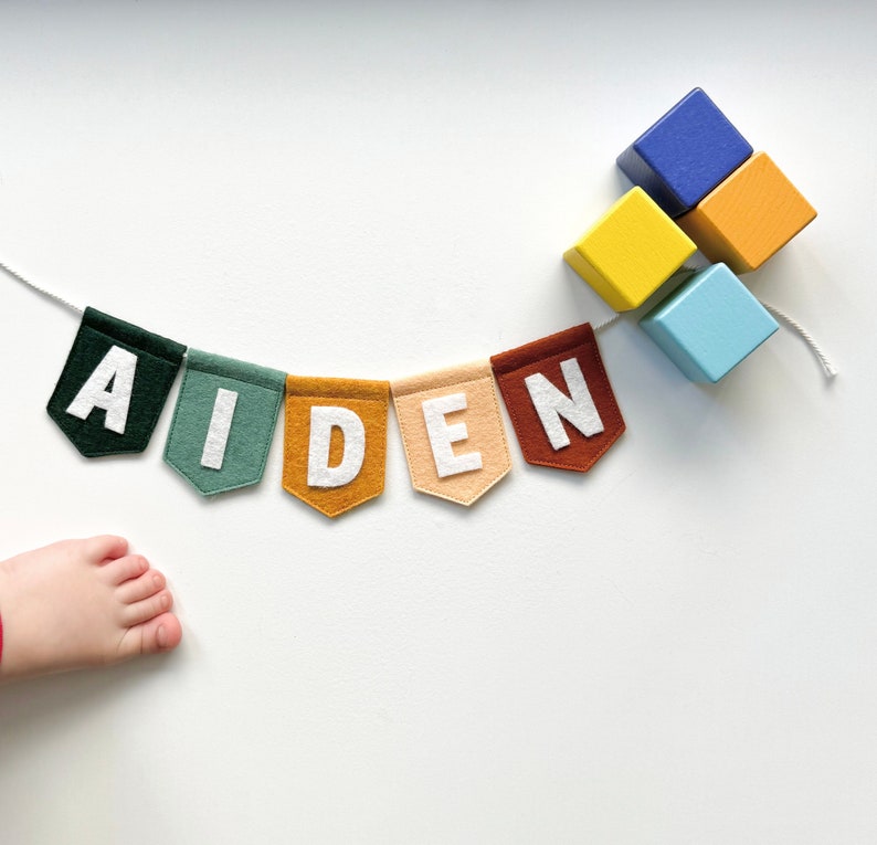 May include: A colorful felt banner spelling "AIDEN" with white letters on various colored pennants. The banner is strung on a white cord, and a cluster of wooden blocks in blue, yellow, and orange are nearby. A child's foot is visible in the corner.
