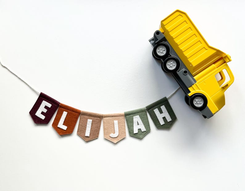 May include: A colorful felt banner spelling "ELIJAH" with white letters, strung on a white cord. The banner is in shades of brown, tan, and green. A yellow toy dump truck rests beside the banner on a white background.