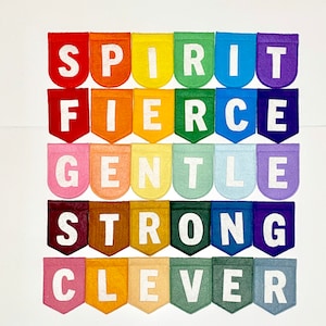 May include: A colorful felt banner with the words "SPIRIT, FIERCE, GENTLE, STRONG, CLEVER" displayed on individual pennants. Each pennant is a different color of the rainbow, with white letters. The banner is arranged in a grid pattern.