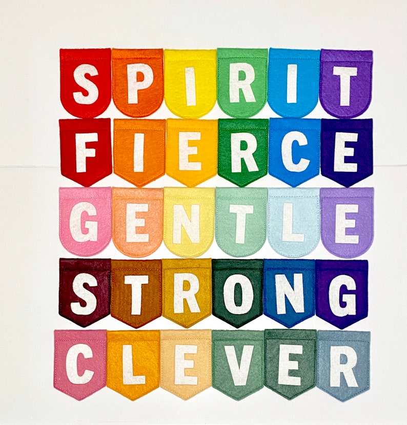 May include: A colorful felt banner with the words "SPIRIT, FIERCE, GENTLE, STRONG, CLEVER" displayed on individual pennants. Each pennant is a different color of the rainbow, with white letters. The banner is arranged in a grid pattern.