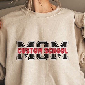 May include: A beige sweatshirt with a black and red graphic that says "MOM CUSTOM SCHOOL" in a varsity style font.