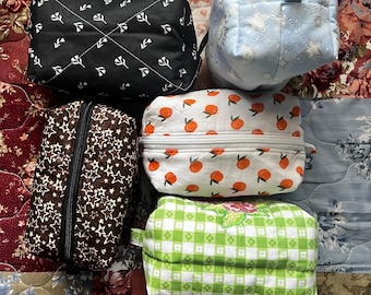 May include: A selection of handmade fabric pouches in diverse patterns and colours. Designs include a black pouch with white floral accents, a light blue pouch with stars, a brown pouch with stars, a white pouch with orange fruit, and a green and white checkered pouch.