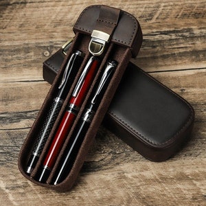 Könnte beinhalten: Ein braunes Leder-Stift-Etui mit Klappenverschluss und einem Metallverschluss. Das Etui enthält drei Stifte, einen mit einer Carbonfaser-Oberfläche, einen mit einer roten Oberfläche und einen mit einer silbernen Oberfläche.