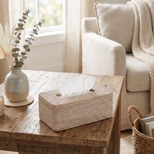 May include: A rectangular tissue box cover crafted from woven light beige rattan material. The box features a clean, structured design with a central rectangular opening at the top, through which a single white paper tissue is pulled upward.